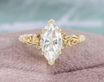 May include: A marquise-cut diamond engagement ring with a gold band. The ring features a large, clear diamond set in a prong setting. The band has intricate filigree detailing on the sides, adding a vintage aesthetic. The ring is displayed on a pink surface.