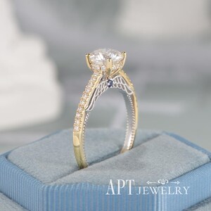 May include: A gold and silver engagement ring with a large, clear center stone. The ring features a detailed design with smaller stones and a blue accent. The ring is displayed in a blue jewelry box. The text "APT JEWELRY" is visible.