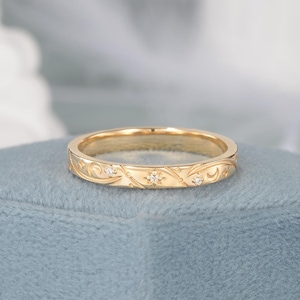 May include: A gold-colored ring with an engraved pattern of stars, swirls, and small diamonds. The ring is set on a blue surface, showcasing its intricate design and craftsmanship. The ring's band is approximately 2 mm wide.
