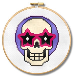 May include: Cross-stitch design of a skull wearing star-shaped sunglasses, framed in a wooden embroidery hoop. The skull is beige, with a purple halo, black teeth, and pink and black sunglasses. The design is on white fabric.