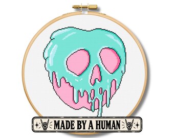 May include: A circular wooden embroidery hoop containing a cross-stitch design of a pink skull covered in dripping mint green liquid. Below the hoop is a rectangular label with the text MADE BY A HUMAN flanked by two hand gesture icons.