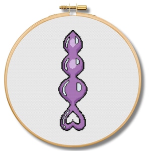 May include: A cross-stitch design of a purple sex toy with a heart-shaped base, displayed in a wooden embroidery hoop. The design features a series of rounded shapes and a pointed top.