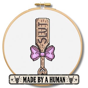 May include: A circular wooden embroidery hoop containing white fabric with a cross-stitch design of a light brown wooden paddle featuring the word SLUT in dark brown letters and a light purple bow. A rectangular label below reads MADE BY A HUMAN.