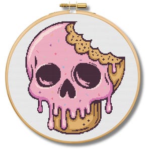May include: A cross-stitch embroidery hoop featuring a pixelated design of a skull covered in pink frosting, with a bite taken out of the top. The skull has dark eye sockets and a dripping frosting effect. The embroidery hoop is round and made of wood.