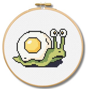 May include: Cross-stitch embroidery of a whimsical snail with an eggshell and yolk shell. The snail's body is green, and the egg is white and yellow. The embroidery is set in a wooden hoop.
