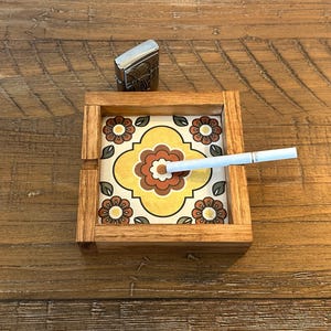 Puede incluir: Cenicero de madera con un inserto de azulejo floral en tonos amarillos, naranjas y marrones. Un cigarrillo encendido descansa en el cenicero. Un encendedor plateado se coloca junto al cenicero sobre una superficie de madera.