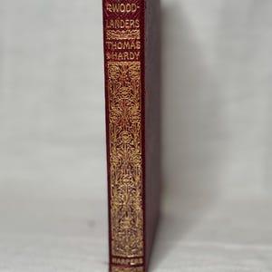 May include: A vintage book titled "The Woodlanders" by Thomas Hardy. The book has a burgundy cover with gold lettering and decorative accents. The spine features the author's name and publisher, "Harpers".