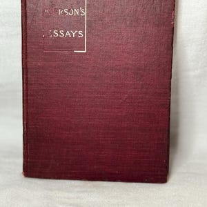 May include: A maroon-coloured hardcover book titled "Emerson's Essays" in a white rectangular frame. The book has a textured cover and is slightly worn, suggesting it is vintage or antique. The book is standing upright.