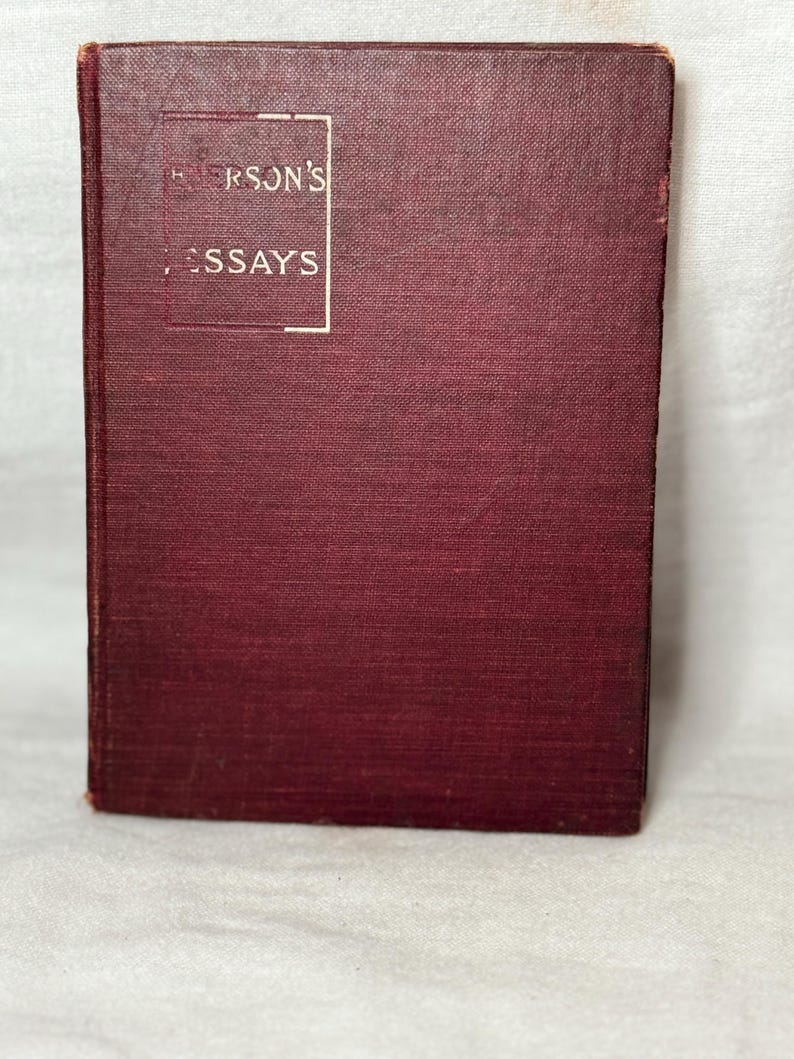 May include: A maroon-coloured hardcover book titled "Emerson's Essays" in a white rectangular frame. The book has a textured cover and is slightly worn, suggesting it is vintage or antique. The book is standing upright.