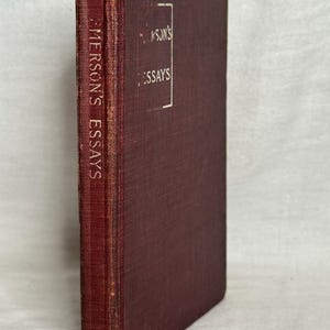 May include: A maroon-coloured hardcover book titled "Emerson's Essays." The spine displays the title vertically, while the front cover features the title within a white rectangular frame. The book has a worn appearance.