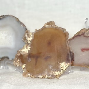 May include: Three agate slices displayed on clear stands. The slices vary in color, with shades of white, brown, and pink. Some edges are trimmed with gold. The agate has a natural, organic appearance, showcasing unique patterns and textures.