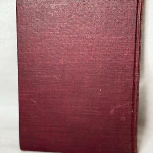 May include: A maroon book with a textured cover. The book is upright, displaying the spine and front cover. The cover has a slightly faded appearance, with minor imperfections. The book's edges are visible, suggesting a vintage or antique item.