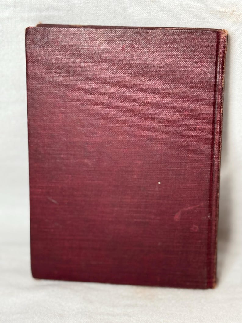May include: A maroon book with a textured cover. The book is upright, displaying the spine and front cover. The cover has a slightly faded appearance, with minor imperfections. The book's edges are visible, suggesting a vintage or antique item.
