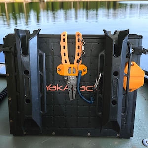 May include: Black fishing rod holder with orange accents, including pliers and a mounting system. The text "YakAttack" is visible on the holder. The background shows a lake and trees.