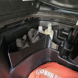 Puede incluir: Un compartimento de almacenamiento de plástico negro en el maletero de un vehículo, que contiene correas negras, dos botellas y una botella de jabón de manos. Una bolsa naranja es visible en la parte inferior. La palabra "RIDGELINE" está impresa en el compartimento.