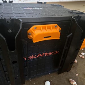 May include: Black storage container with an orange handle and the text "YakAttack" in red. The container has a textured surface and is on a sandy beach with seashells. The container is designed for outdoor use.