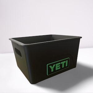Puede incluir: Contenedor de almacenamiento rectangular negro con el logotipo "YETI" verde. El contenedor tiene un recorte de asa en el lateral y un borde ligeramente elevado. Fabricado con material duradero.