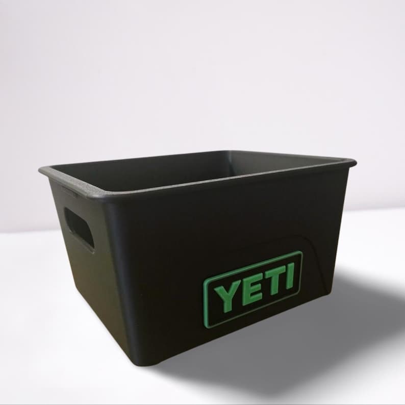Puede incluir: Contenedor de almacenamiento rectangular negro con el logotipo "YETI" verde. El contenedor tiene un recorte de asa en el lateral y un borde ligeramente elevado. Fabricado con material duradero.