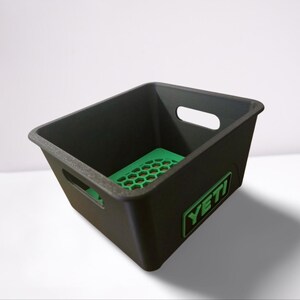 Puede incluir: Contenedor de almacenamiento de pl&aacute;stico negro con una base verde con patr&oacute;n de panal. El contenedor tiene dos recortes para asas y un logotipo verde "YETI".