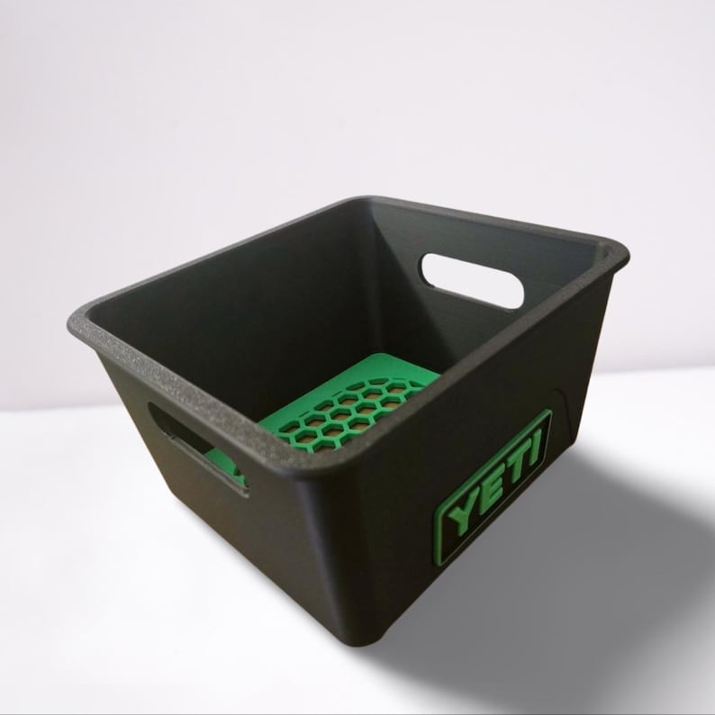 Puede incluir: Contenedor de almacenamiento de pl&aacute;stico negro con una base verde con patr&oacute;n de panal. El contenedor tiene dos recortes para asas y un logotipo verde "YETI".