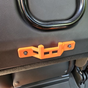 May include: An orange 3D-printed bracket, secured with black screws, is mounted on a black textured surface. The bracket has a geometric design with rectangular cutouts and is positioned near a black metal bar.