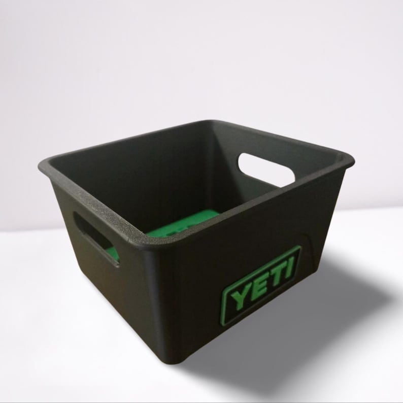 Puede incluir: Un contenedor de almacenamiento cuadrado negro con una base interior verde y dos recortes de asa. El contenedor tiene la palabra "YETI" en verde en la parte delantera. El contenedor est&aacute; hecho de un material duradero.