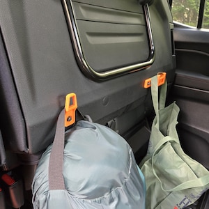 Könnte beinhalten: Autoinnenraum mit schwarzer Sitzlehne und silberfarbenem Metallgriff. Orangefarbene Haken halten einen grauen Schlafsack und eine grüne Tragetasche. Die Taschen hängen im Heck des Fahrzeugs.
