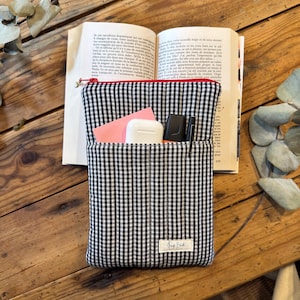 Peut inclure: Une pochette matelassée en tissu vichy noir et blanc avec une fermeture éclair rouge. La pochette comporte une poche avant contenant un post-it rose, un boîtier d'écouteurs sans fil blanc, un surligneur orange et un stylo noir. Une étiquette blanche est cousue.