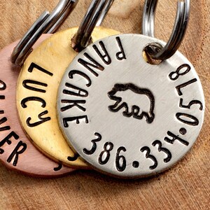 May include: Three round metal dog tags with engraved names and phone numbers. The tags are attached to a silver key ring. The tags are gold, copper, and silver. The silver tag has a small bear silhouette engraved on it.