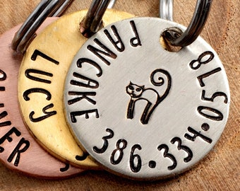 Op de afbeelding: Drie ronde metalen huisdier-ID-tags met sleutelringen. De tags zijn in verschillende kleuren: koper, goud en zilver. De koperen tag zegt "OLIVER", de gouden tag zegt "LUCY" en de zilveren tag zegt "PANCAKE 386" met een klein katten silhouet en het telefoonnummer "334-0578".