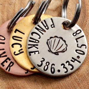 May include: Three personalized pet ID tags with names and contact information. The tags are made of copper, gold, and silver-colored metal. One tag features a seashell design. Each tag has a metal ring for attachment.