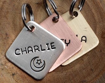 May include: Three metal pet tags with names and a crescent moon and star symbol. The tags are silver, copper, and gold-colored, each with a silver ring for attachment. The tag for "Charlie" is silver.