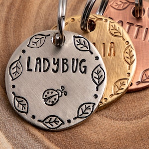 May include: Three round metal pet tags with leaf designs and engraved names. The tags are silver, gold, and copper. The silver tag reads "Ladybug" and has a ladybug design. The gold tag reads "Mia" and the copper tag reads "Lottie".