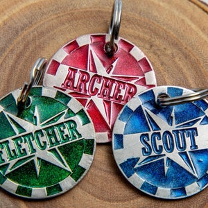 Puede incluir: Tres chapas redondas de metal para perros con diseños de estrellas y bordes blancos. Las chapas son roja, verde y azul con los nombres "Archer", "Fletcher" y "Scout" escritos en ellas.