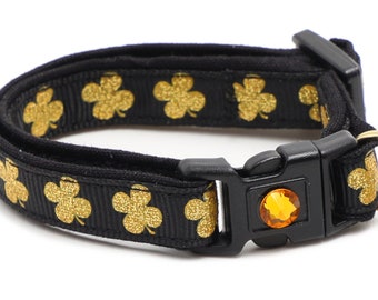 May include: Black cat collar with a gold shamrock pattern and a small bell. The collar has a clear plastic buckle with a gold-colored gem.