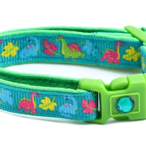 May include: A green cat collar with a bell and a turquoise ribbon featuring a dinosaur pattern.