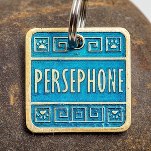 Square pet tag with rounded corners. The tag is gold and light blue. The example name reads Persephone. It is large and in all uppercase letters across the center. Above and below the name is a border of Greek key flourishes and paw prints.