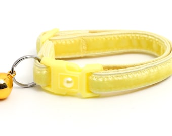 Könnte beinhalten: Ein hellgelbes Katzenhalsband mit einer goldfarbenen Glocke. Das Halsband besteht aus einem weichen, samtigen Material und verfügt über eine Kunststoffschnalle mit einem perlenartigen Akzent. Die Glocke ist mit einem silberfarbenen Ring befestigt.