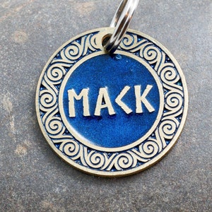 May include: A blue and gold dog tag with the name "MACK" engraved on it. The tag has a decorative border with a swirling pattern.
