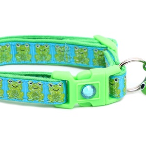 Puede incluir: Un collar para gato verde y azul con un diseño de rana y una campana. El collar tiene una hebilla verde y una gema transparente.