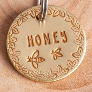 Gold colored round pet tag with darker gold text and design. There are flowers around the edge. The name is straight across the middle. Two Cute bees are under the name.