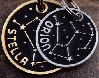 May include: Two silver dog tags with black backgrounds and gold lettering. The top tag says "Orion" and features a constellation design. The bottom tag says "Stella" and also features a constellation design.
