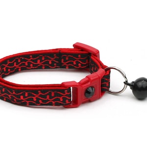 May include: Red and black cat collar with a breakaway buckle and a black bell. The collar has a wavy pattern.
