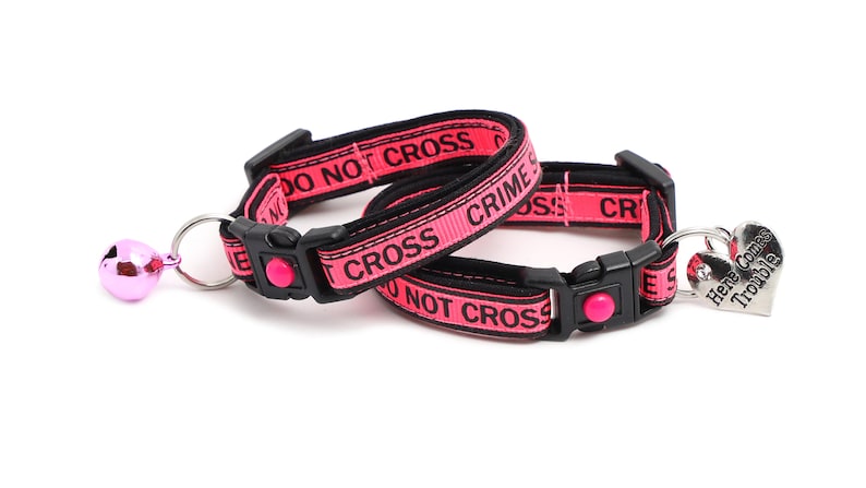 May include: Two pink cat collars with black text that reads "Do Not Cross Crime Scene" and a silver heart-shaped tag that reads "Here Comes Trouble".