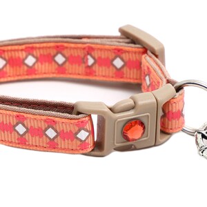 Puede incluir: Un collar para gato con estampado naranja y rosa con un dije plateado y una campanita rosa.