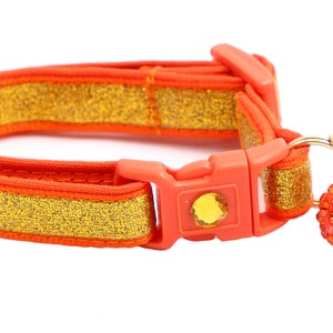 Op de afbeelding: Oranje en gouden kattenhalsband met een bel en een glinsterende hanger. De halsband heeft een plastic gesp.