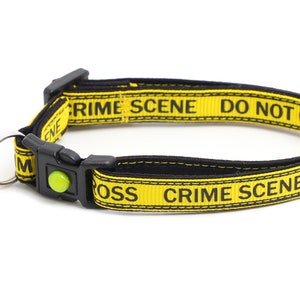 Op de afbeelding: Gele en zwarte kattenhalsband met een bel. De halsband heeft de tekst "Crime Scene Do Not Cross" erop gedrukt.
