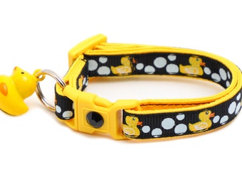 Puede incluir: Un collar para gato amarillo y negro con un dije de pato de goma. El collar tiene un fondo negro con círculos blancos y patos de goma amarillos impresos en él.