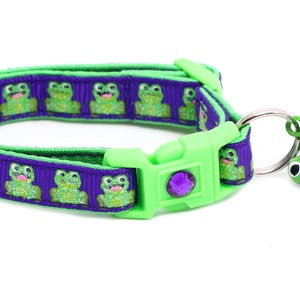 Puede incluir: Un collar para gato morado y verde con estampado de rana y una campana verde con forma de rana. El collar tiene una hebilla de plástico y una gema morada.