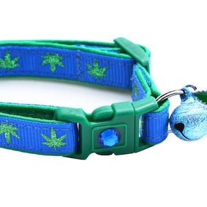 May include: Blue and green cat collar with a cannabis leaf pattern. The collar has a green buckle with a blue gem, a blue bell, and a green cannabis leaf charm.